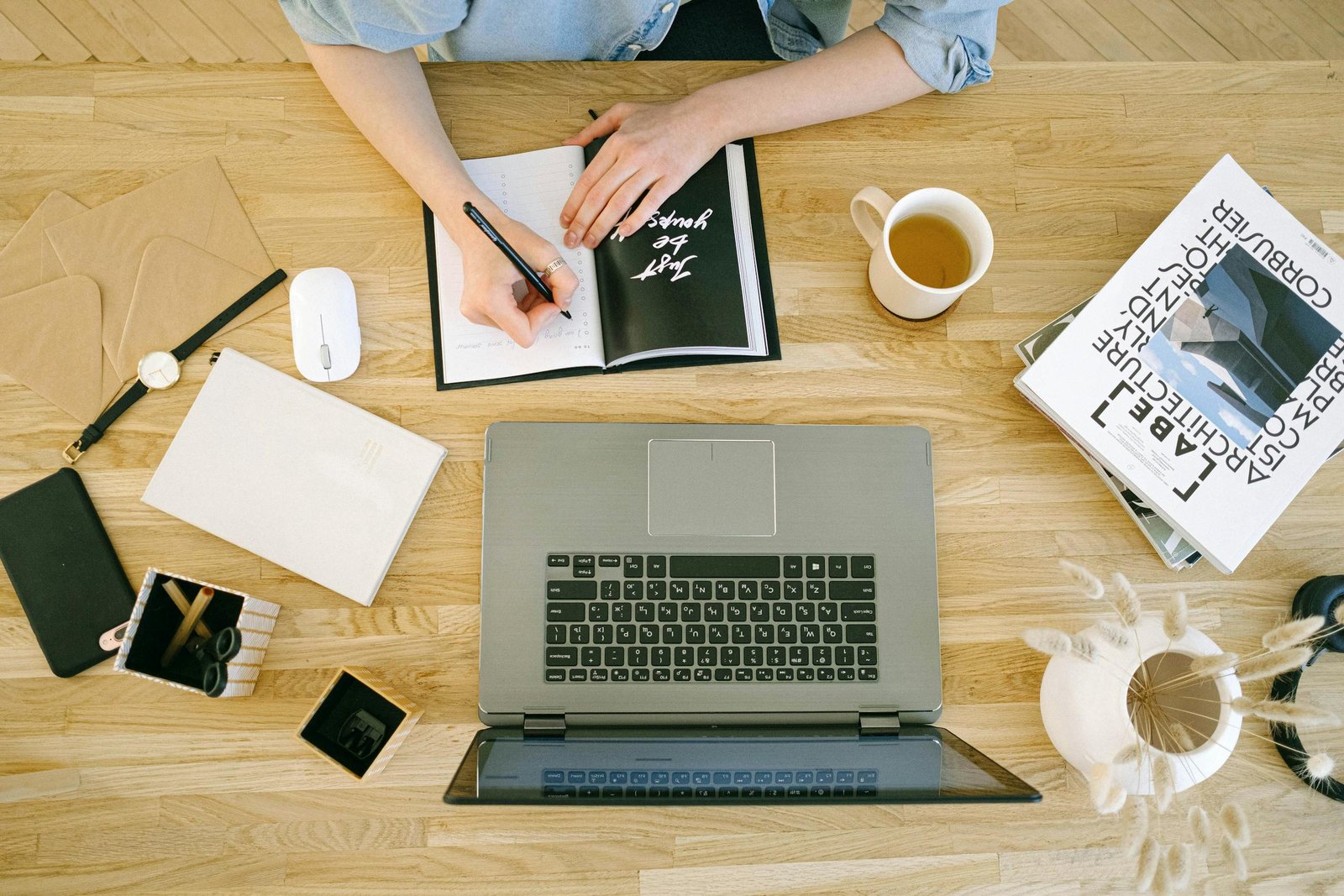 A woman writing notes at a home office desk with a laptop and coffee cup.